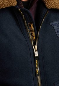 Námořnická vlněná bunda s krémovým umělým beránkem, zlatým zipem a brandingem PME Legend. Na přední části je nášivka s motivem orla.