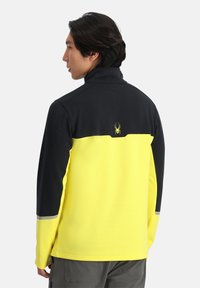 Pullover in pile con colletto alto, parte superiore nera e parte inferiore gialla brillante, con logo a forma di ragno e dettagli riflettenti.
