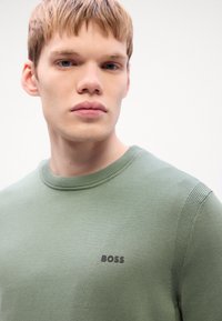 Green knit sweater with a crew neck. Features subtle ribbed accents on shoulders and black "BOSS" logo on the chest. Smooth texture.