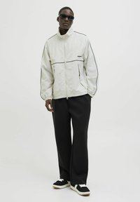 Hvid zip-up jakke med sorte detaljer, løs pasform, stylet med sorte bukser og sorte sneakers med hvide såler. Modellen har mørke solbriller på.