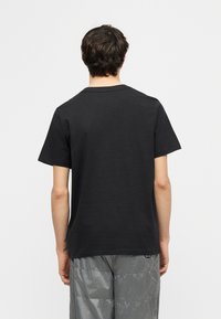 Άτομο με κοντά σκούρα μαλλιά που φοράει μια απλή μαύρη κοντομάνικη μπλούζα και γκρι παντελόνι, όρθιο με την πλάτη προς τα εμπρός σε λευκό φόντο.