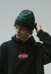 Strickmütze in Schwarz und Türkis mit einem abstrakten Muster; hat einen umgeschlagenen Bund und ein lila Logo-Patch. Wird mit einem schwarzen Hoodie getragen.