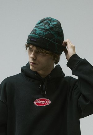 Knit beanie in black and teal with an abstract pattern; features a folded cuff and a purple logo patch. Worn with a black hoodie.
