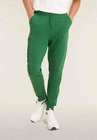 Pantaloni sportivi verdi con design affusolato, vita elastica, tasche laterali e un dettaglio del logo testurizzato lungo la gamba. Abbinati a sneakers bianche.