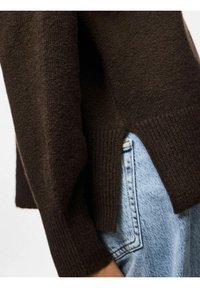 Maglione marrone lavorato a maglia con polsini a costine e spacchi laterali, abbinato a jeans blu chiaro. LaTexture appare morbida e leggermente pelosa.