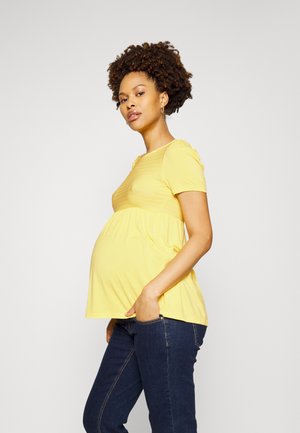 Top de maternidade amarelo de manga curta com textura franzida sob o busto, combinado com jeans azul escuro. O modelo está de perfil.