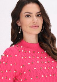 Pulover roz tricotat cu un pattern texturat, embellisat cu rânduri de margele albe și perle de aur. Modelul are părul lung, ondulat, și cercei rotunzi cu mărgele.