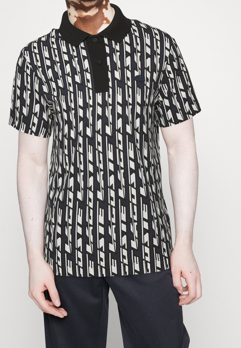 Personne portant un polo à motifs géométriques noirs et blancs avec un col noir et un pantalon foncé, sur un fond uni.