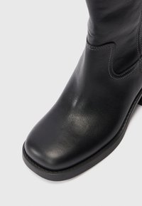 Botín de cuero negro que presenta una puntera redondeada, textura suave, costuras mínimas y un tacón elevado con una suela de goma.