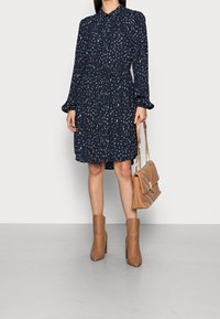 Donna che indossa un vestito a maniche lunghe blu navy con pois, stivali alla caviglia color sabbia e tiene una borsa a tracolla trapuntata coordinata in tonalità sabbia con catena dorata.