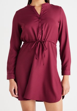 Vrouw draagt een bordeauxrode jurk met lange mouwen, een knoopsluiting bij de hals en een koord in de taille.