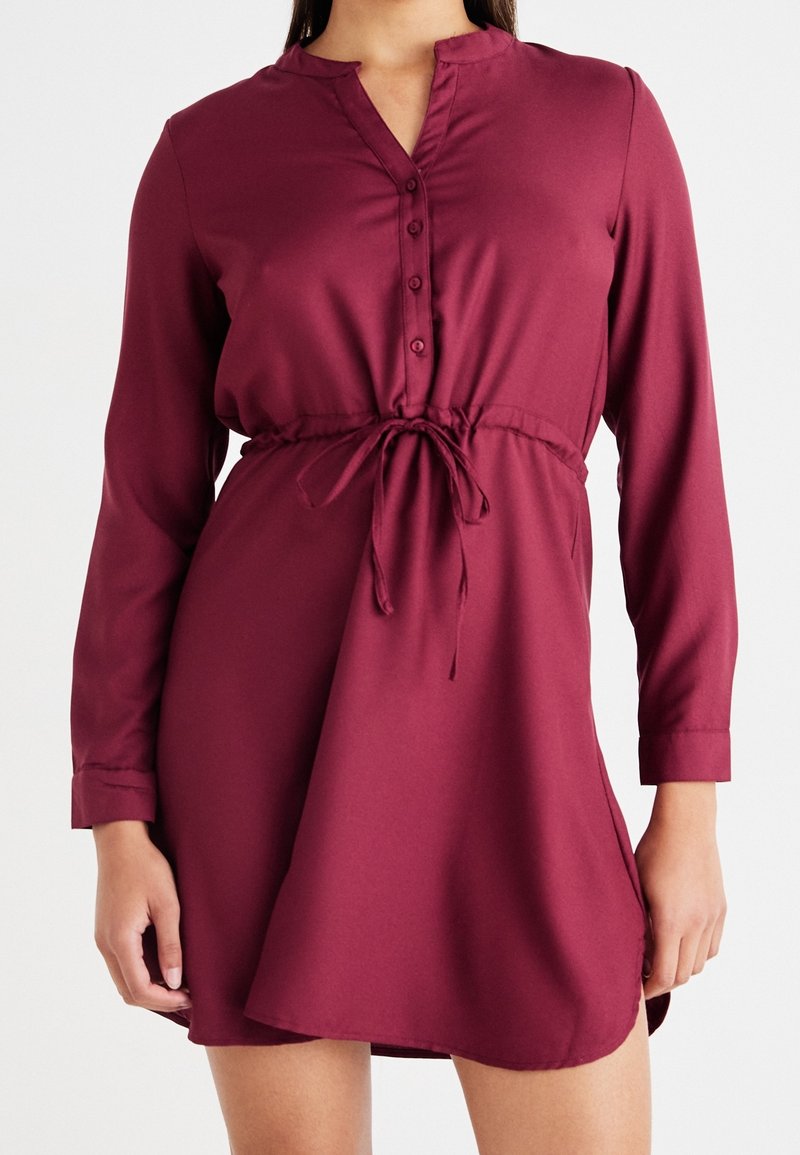 Femme portant une robe bordeaux à manches longues avec un col boutonné et un cordon noué à la taille.