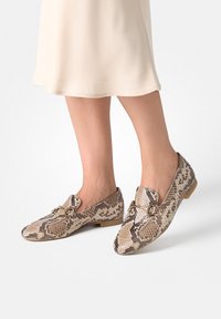 Nohy oblečené v béžové sukni a mokasíny se vzorem hadí kůže se zlatým detailem ve tvaru koníka na bílém pozadí.