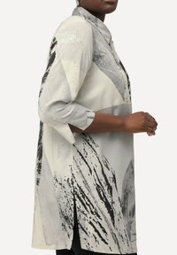 Blusa a maniche lunghe color grigio chiaro con motivi astratti neri; presenta un colletto a punta e fessure laterali per un comfort aggiuntivo.