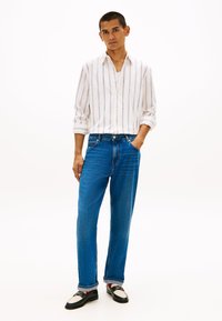 Άσπρο πουκάμισο με κουμπιά και κάθετες ρίγες, συνδυασμένο με φαρδιά μπλε τζιν και μαύρα παπούτσια με λευκές λεπτομέρειες. Το ύφασμα φαίνεται λείο και άνετο.