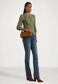 Mulher vestindo um cardigan verde de malha trançada, jeans azuis, botas de salto castanhas e carregando uma bolsa de ombro castanha, de pé contra um fundo branco.