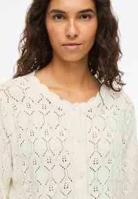 Femme aux cheveux bouclés foncés portant un cardigan en maille crème avec un col festonné et des boutons en perles, face à l'avant sur un fond blanc.