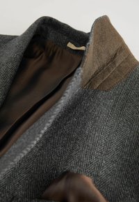 Innenansicht eines grauen Fischgrät-Sakkos mit braunem Satinfutter, auffälligem Kragen in Braun und maßgeschneiderten Steppnähten.