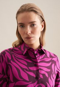 Koszula z wyrazistym wzorem liści w kolorze fioletowym i różowym, klasycznym kołnierzykiem oraz czarnymi guzikami, wykonana z gładkiego materiału o lekko błyszczącej powierzchni.