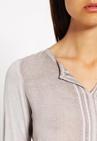 Blusa grigia chiara con scollo a V, maniche lunghe e una trama sottile. Presenta un colletto a pieghe e plissettature verticali sul davanti.