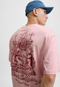 Lyserød bomulds T-shirt med et rødt grafisk design med engle og lysekroner. Teksten lyder "Tid er penge", korte ærmer, afslappet pasform.