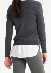 Maglione grigio carbone lavorato a maglia con maniche lunghe, orlo a coste, indossato sopra una camicetta bianca a righe, abbinato a pantaloni grigi.