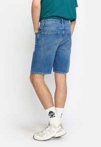 Lichte blauwe denimshorts met een ontspannen pasvorm, voorzien van achterzakken en omgeslagen randen. Gecombineerd met witte sokken en grijze sneakers.