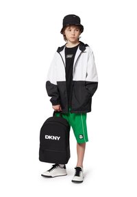 Veste coupe-vent noir et blanc, chapeau seau noir, sac à dos noir avec le logo "DKNY", shorts verts et baskets blanches.