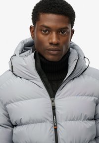 Giacca puffer grigia chiara con cappuccio, caratterizzata da cuciture orizzontali, zip frontale e un maglione nero sotto. Logo visibile sulla zip.
