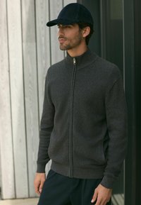 Dunkelgrauer Reißverschluss-Sweatshirt mit strukturierter Oberfläche und hohem Kragen, kombiniert mit einer marineblauen Cap und schwarzen Hosen, vor einem Holz-Hintergrund stehend.