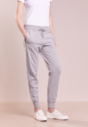 Persoon draagt lichtgrijze joggingbroek met trekkoord, wit T-shirt met korte mouwen en witte sneakers, handen in de zakken, staand op een beige vloer.