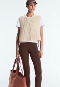 Femeie purtând o vestă matlasată bej peste un tricou alb și colanți maro, ținând o geantă mare maro, purtând o șapcă maro, uitându-se spre dreapta.