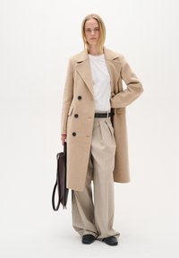 Bēšas vilnas maisījuma mēteles ar lieliem melniem pogām, valkāta virs baltas T-kreklu un pleatotām bēšām biksēm. Melni apavi un tumši brūna soma.