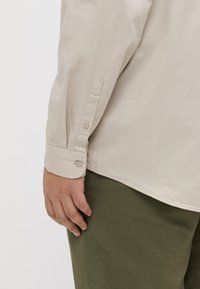 Camisa beige de manga larga con puños abotonados, acompañada de pantalones verde oliva, que presentan una textura suave y un corte relajado.