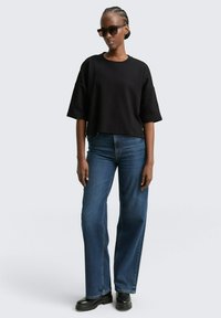 T-shirt noir court avec des manches larges, associé à un jean bleu taille haute. Les accessoires comprennent des lunettes de soleil rectangulaires sombres et des bottes noires.