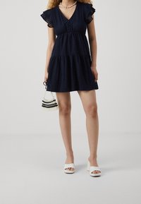 Granatowa sukienka koronkowa z falbanami na rękawach, wielowarstwowym fasonem i dekoltem w serek. Do tego białe sandały na grubym obcasie i torebka z koralików.