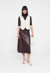Chaleco blanco con botones sobre un cuello de tortuga negro, combinado con una falda de cuero burdeos y tacones abiertos color crema, sosteniendo un bolso blanco.