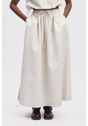 Kobieta ubrana w kremową, wysokowalistą spódnicę maxi z troczkiem i rozcięciami po bokach, w połączeniu z ciemnobrązowymi botkami za kostkę.