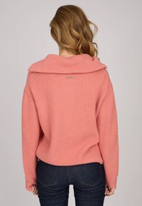 Pull en tricot rose à côtes avec un large col drapé et des manches longues, doté d'une petite étiquette métallique au dos, associé à un jean sombre.