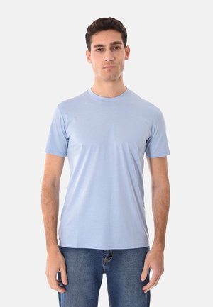 Uomo in piedi di fronte che indossa una maglietta a maniche corte azzurro chiaro e jeans blu su uno sfondo bianco uniforme.