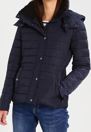 Woman wearing a navy blue quilted puffer jacket with a hood, snap buttons, and front pockets, paired with blue jeans.
