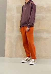 Lila hoodie med framficka och broderad logotyp, matchad med orangea sweatpants med sidologotyp, kompletterad med svarta och vita randiga skor.