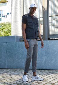 Juodas liemenė su daugybe kišenių, vilkima virš prigludusios juodos marškinėlių, derinama su pilkų kelnių ir baltų sportbačių. Lauko miesto aplinka.