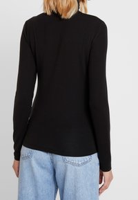 Persona che indossa un top aderente nero a maniche lunghe e jeans in denim blu chiaro, mostrata di spalle su uno sfondo bianco semplice.