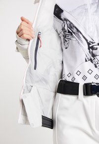 Personne en train de dézipper une veste blanche révélant une poche intérieure zippée, portant un pantalon blanc avec une ceinture noire et un haut imprimé graphique.