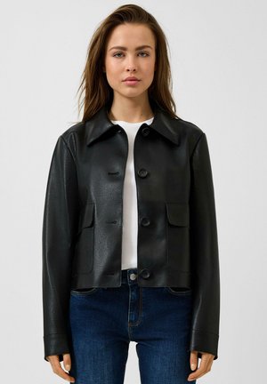 Junge Frau trägt eine schwarze, geknöpfte Kunstlederjacke über einem weißen Top und blauen Jeans, steht vor einem schlichten, hellen Hintergrund.