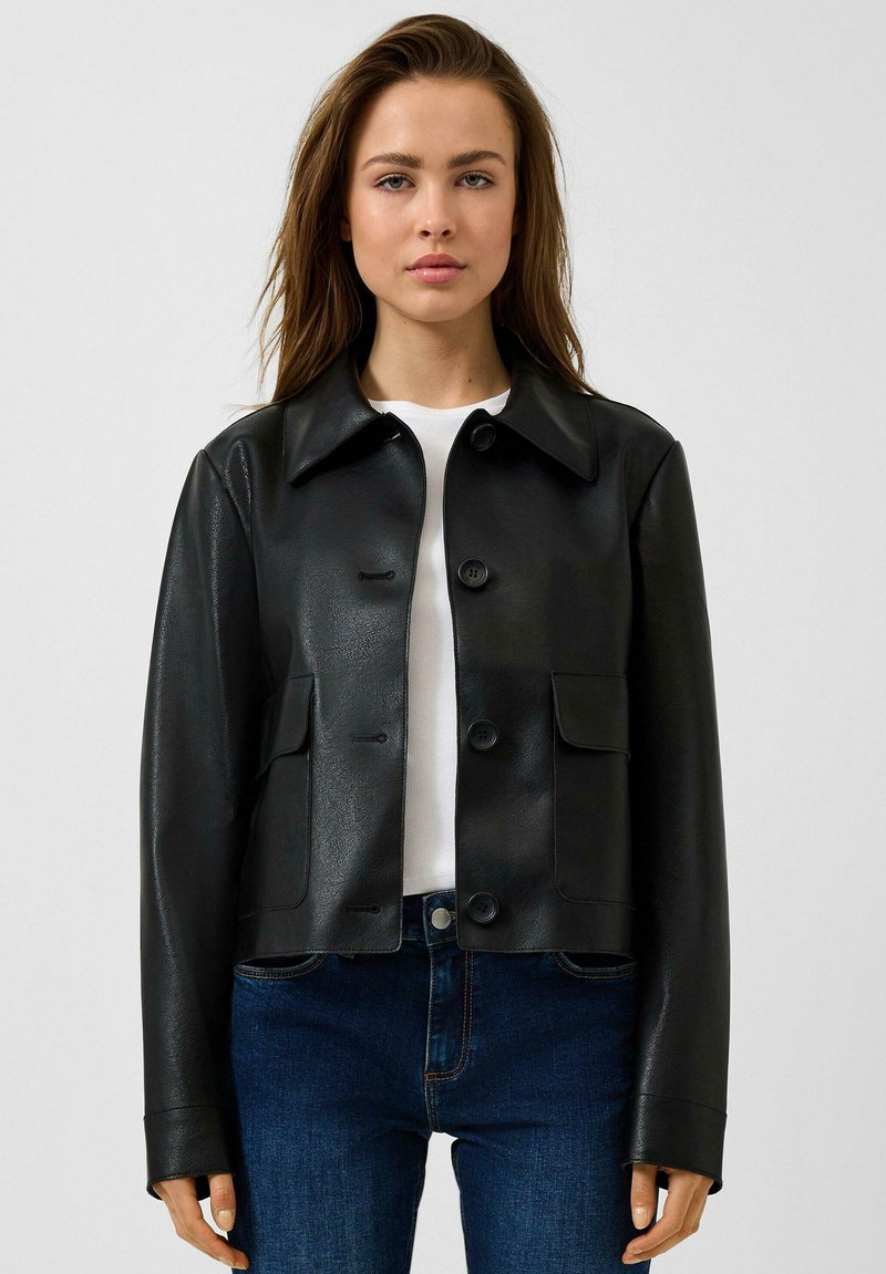 Jonge vrouw die een zwarte, met knopen gesloten, nep leren jas draagt over een wit shirt en blauwe spijkerbroek, staand tegen een effen lichte achtergrond.