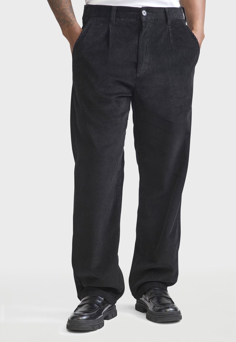 Pantalons en velours côtelé noir à coupe droite, avec des plis avant, des poches latérales et une fermeture à bouton, associés à des chaussures noires.