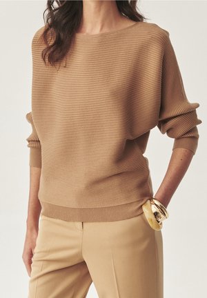 Vrouw met een geribde trui in kamelkleur en bijpassende broek, met gouden en beige armbanden om haar rechterpols, hand in de zak.
