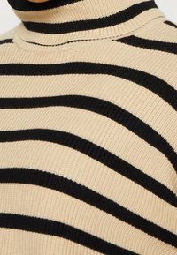 Pull en col roulé beige avec des rayures horizontales noires. Texture côtelée, design ajusté et détail de col haut visible.